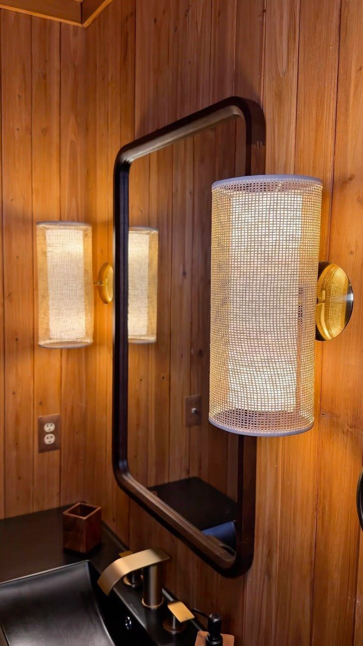 Cylindrical mesh sconces flanking a black-framed mirror on a wood-paneled bathroom wall.